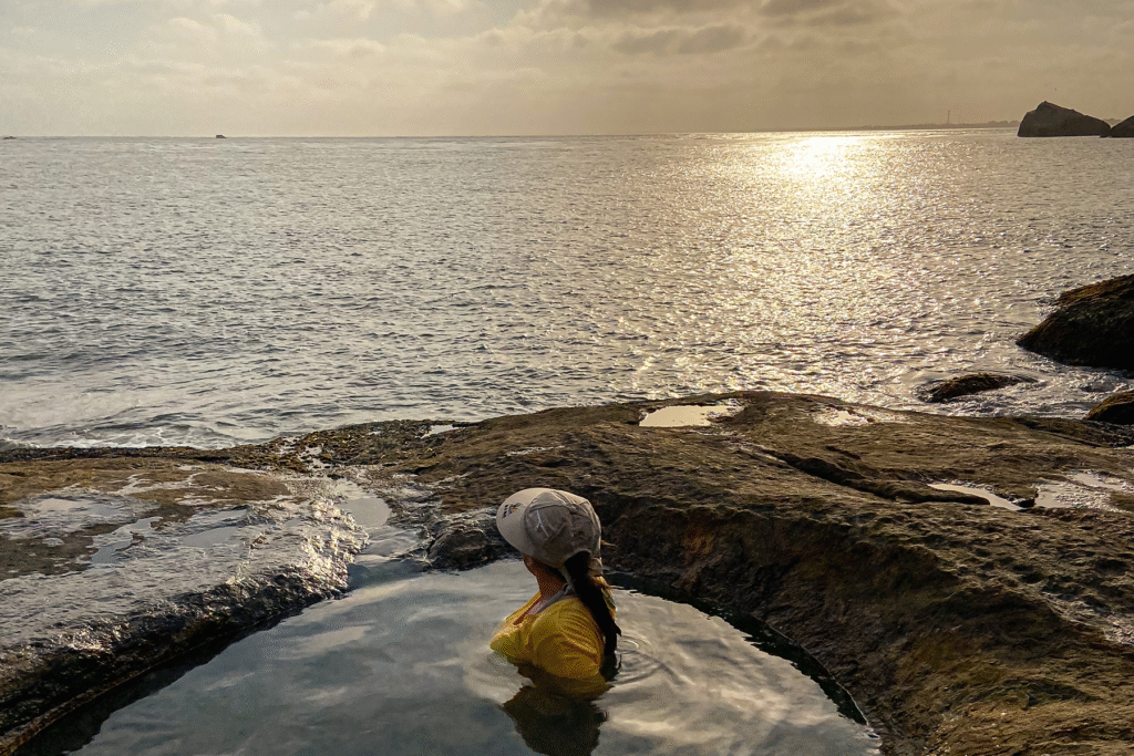 Uma mulher de costas, em um buraco da pedra cheio de água, com o mar ao fundo e o pôr do sol