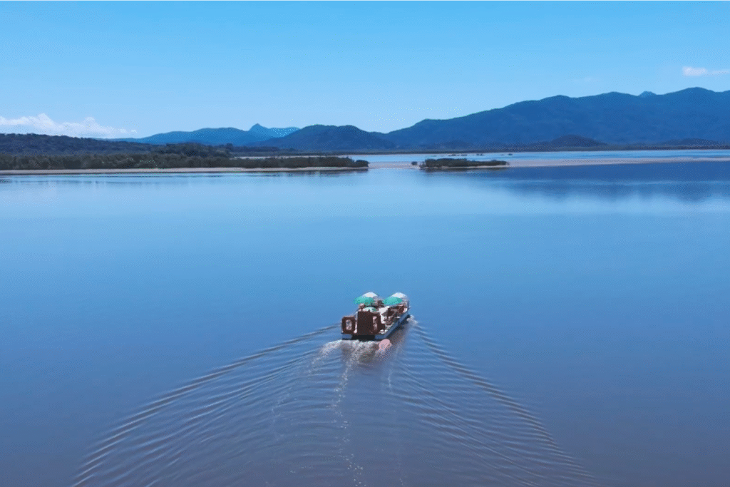 imagem aérea da baía de antonina, enorme, um veradeira espelho de água, com um barquinho pequeno no meio. a água reflete o ceú azul, e os morros ao fundo cortam a foto no meio